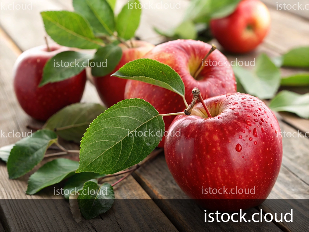 A close-up of glistening red apples accompanied by green leaves, resting on a rustic wooden table. A few apples are positioned in the background, enhancing the natural, fresh look.