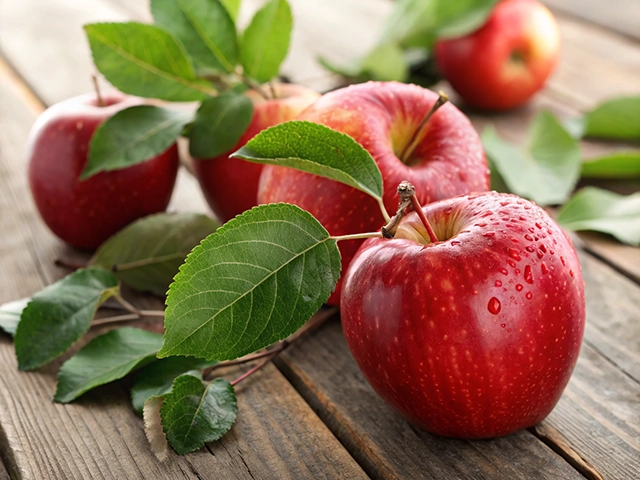 A close-up of glistening red apples accompanied by green leaves, resting on a rustic wooden table. A few apples are positioned in the background, enhancing the natural, fresh look.
