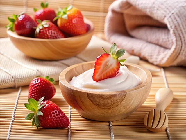A wooden bowl filled with creamy yogurt, topped with a fresh strawberry, accompanied by additional strawberries in another bowl, set against a bamboo mat background.