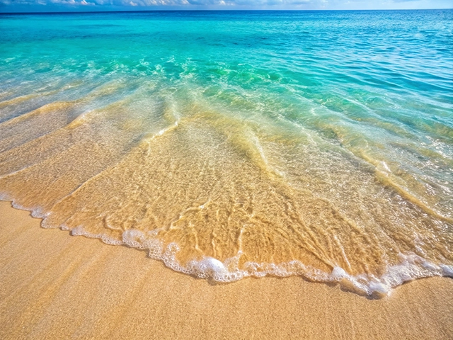 A serene beach scene featuring gentle waves lapping at a sandy shore. The water is a gradient of turquoise and blue, contrasting beautifully with the golden sand. The horizon is clear with a few clouds, creating a tranquil atmosphere.